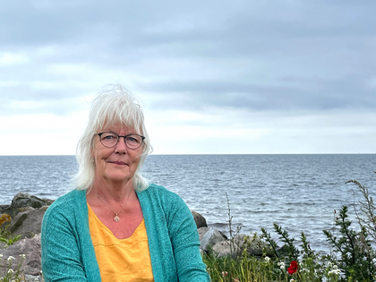 Äldre kvinna med vitt hår och glasögon sitter vid havet, klädd i gul tröja och turkos kofta.