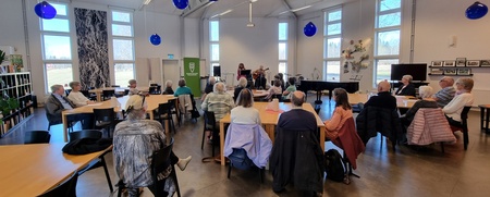 Ett antal personer sitter vid runda bord i en ljus samlingssal med stora fönster och blå lampor i taket.