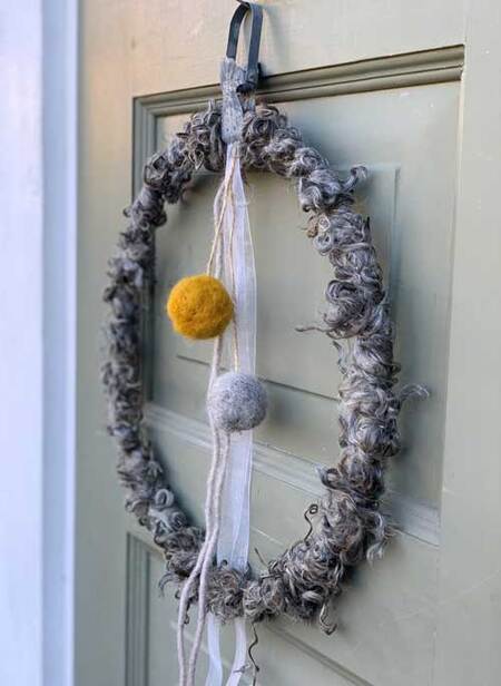 En dörrkrans av grå ull med två tovade bollar, en gul och en grå, samt vita band, hänger på en grön dörr.
