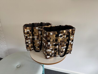 Två väskor flätade av återvunna kaffeförpackningar står på ett runt bord mot en vit vägg.