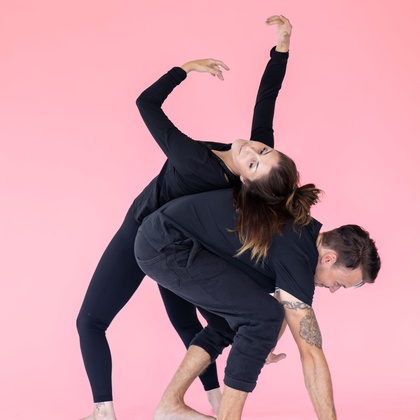 Två personer i svarta kläder utför en modern danspose mot en rosa bakgrund.