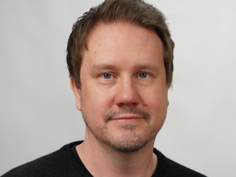 Man med kort brunt hår och skäggstubb, bär en mörk tröja och tittar mot kameran, ljus bakgrund.