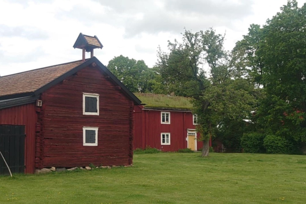Två röda trähus på en grön gräsmatta med träd och molnig himmel i bakgrunden.