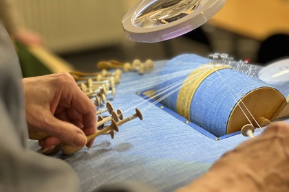 Närbild av händer som tillverkar spets med hjälp av knyppelpinnar och en blå knyppeldyna.