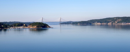 Utsikt över en lugn fjord med en bro i bakgrunden och skogsklädda kullar på sidorna.