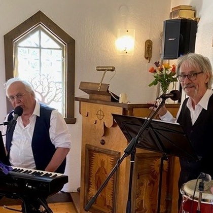 Två musiker spelar i en liten kyrka, en på keyboard och en på congas, båda i vita skjortor och svarta västar.