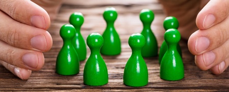 Två händer skyddar en grupp gröna spelpjäser på ett träbord.