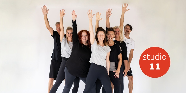 En grupp glada vuxna i träningskläder poserar med armarna uppsträckta framför en vit bakgrund.