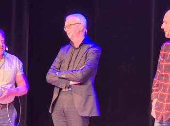 Tre män står på en scen, två i vardagskläder och en i kostym, med mikrofoner och en kopp.