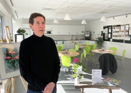 En man i svart tröja står i ett ljust, modernt rum med gröna stolar, runda bord och växter.