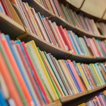 En man står i ett bibliotek och läser i en bok framför en hylla full av färgglada böcker.