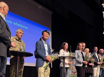 En grupp personer står på en scen med mikrofoner och blommor, framför en projicerad presentation.