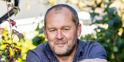 Man sitter utomhus i en trädgård med gröna växter och buskar i bakgrunden.