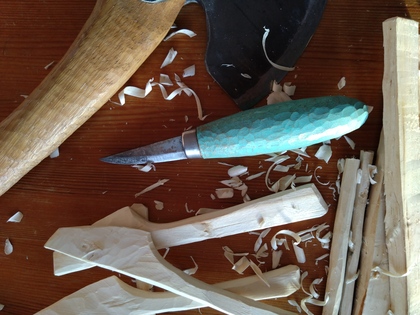 En närbild av träslöjdsarbete med täljda träbitar, en kniv med turkost handtag, en yxa och träspån på ett träbord.