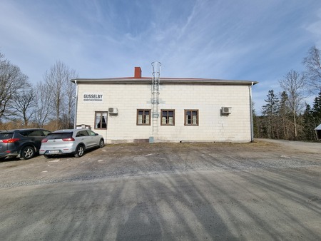 Vit byggnad med skylten 'Gusselby Hembygdsförening', tre bilar parkerade framför, omgiven av träd.