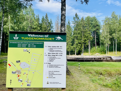 Informationsskylt vid Tuggenområdet i en grön skogsmiljö med blå himmel i bakgrunden.