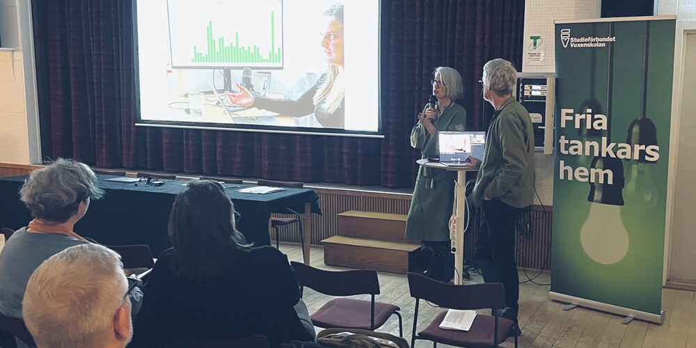 Två personer håller en presentation inför en liten publik med en projektor och en rollup med texten 'Fria tankars hem'.