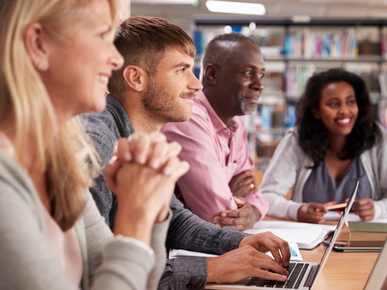 Fyra vuxna personer sitter vid ett bord i ett bibliotek och arbetar tillsammans med datorer och anteckningar.