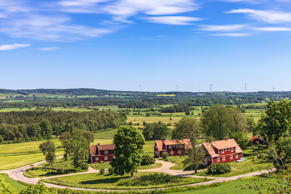 Landskapsvy med röda trähus, gröna fält, träd och vindkraftverk under blå himmel.