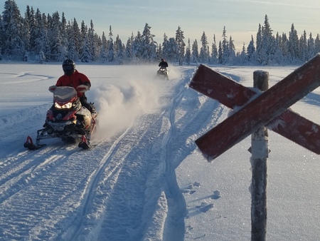 Två personer kör snöskoter på ett snötäckt fält med skog i bakgrunden och en röd krysskylt i förgrunden.