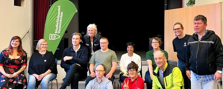 Gruppfoto av tretton personer i olika åldrar, sittande och stående inomhus framför en scen.