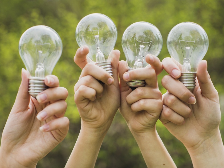 Fyra händer håller upp varsin glödlampa utomhus med grönskande bakgrund.
