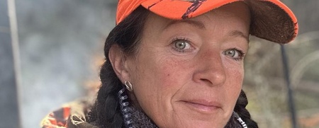 Kvinna med flätor och kamouflagekeps i skogsmiljö, klädd i orange och grönt ytterplagg.