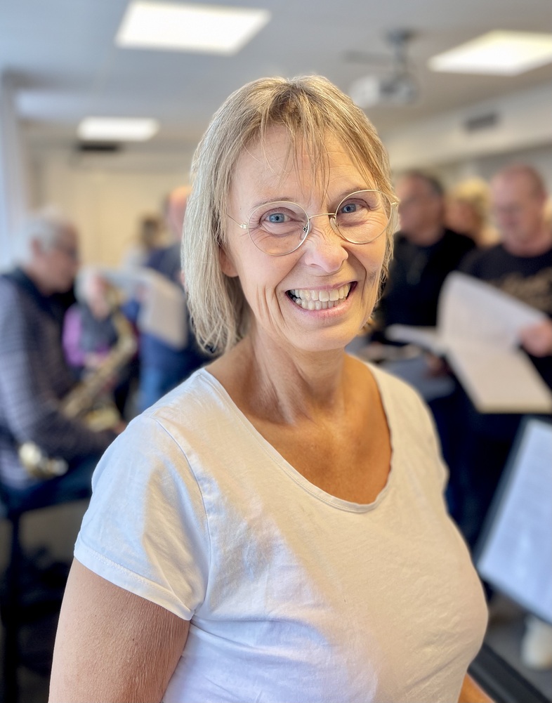 Leende kvinna med glasögon i vit t-shirt står i ett ljust rum med flera personer i bakgrunden.