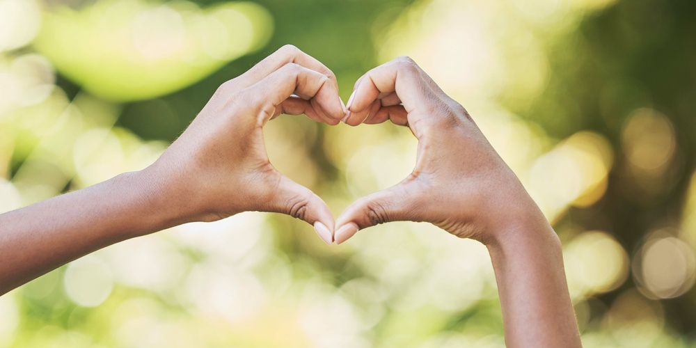 Två händer formar ett hjärta mot en suddig, grön bakgrund utomhus.