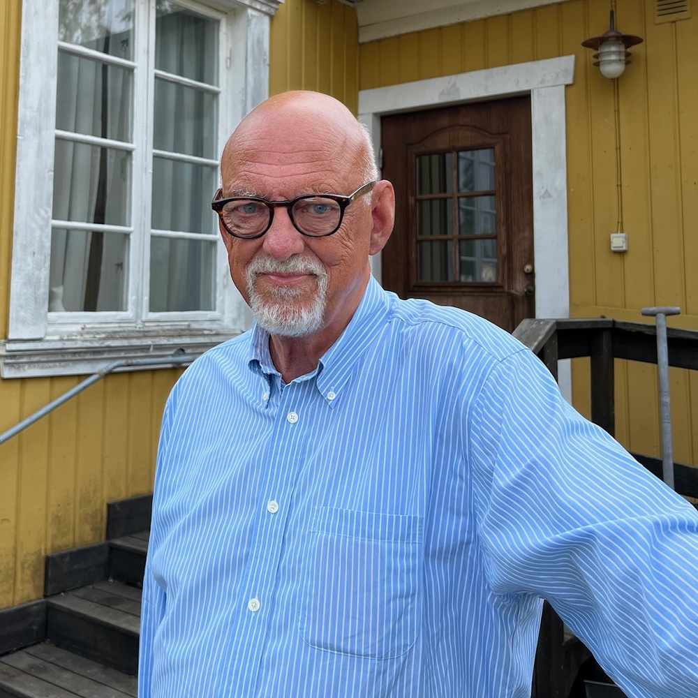 Äldre man med glasögon och blå randig skjorta står utomhus framför ett gult trähus med vita fönsterkarmar.