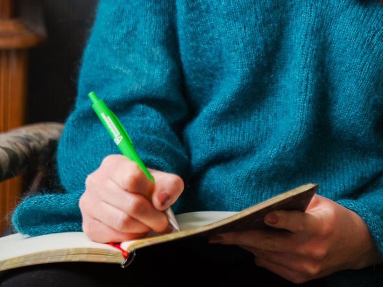 En ung person med långt brunt hår sitter i en stol och skriver i en anteckningsbok med en grön penna.