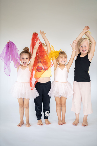 Fyra glada barn står tillsammans, tre i balettkläder och en i svarta byxor, med färgglada sjalar och armarna i luften.