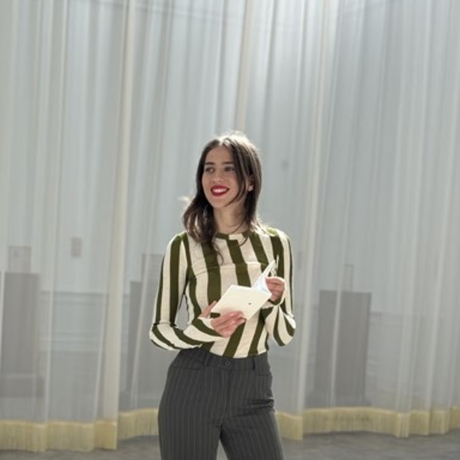 Person i randig tröja och svarta byxor står i ett ljust rum med gardiner och håller ett papper.