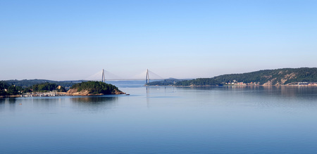 Utsikt över en lugn fjord med en bro i bakgrunden och skogsklädda kullar på sidorna.