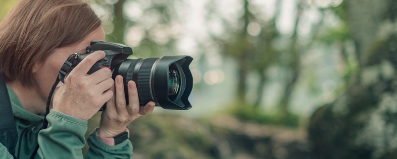 Person med kamera fotograferar i en skogsmiljö, klädd i grön jacka och ryggsäck.