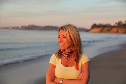 Kvinna med ljust hår står leende på en strand i solnedgången, med armarna i kors.