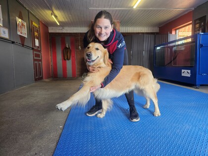 En kvinna ler och håller en glad golden retriever på ett blått golv i ett inomhusutrymme.