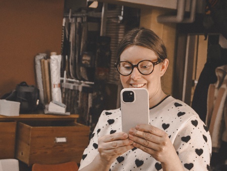 Person med ljust hår håller en mobiltelefon i ett rum med träbyråer och förvaringslådor.