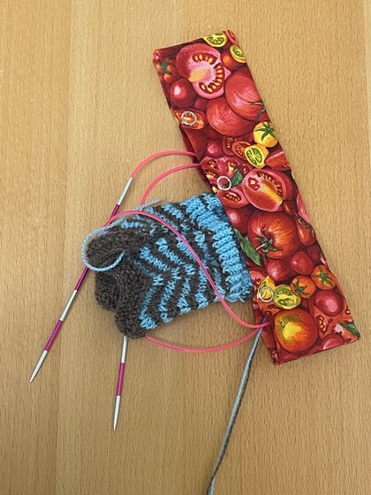 Stickning med blått och brunt garn på rundstickor, bredvid ett fodral med tomatmönster på ett träbord.