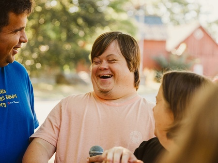 En man med Downs syndrom skrattar tillsammans med två andra personer utomhus en solig dag.