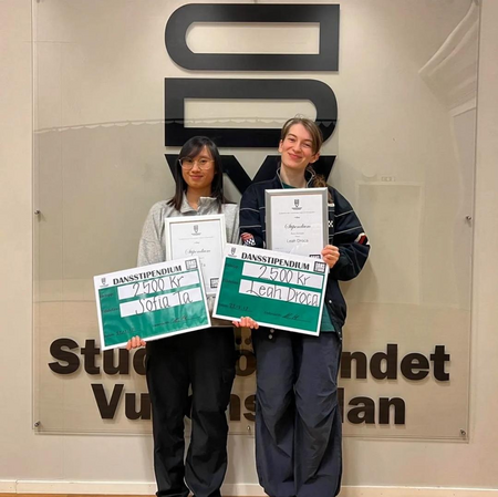 Två unga personer står framför en vägg med logotyp och håller diplom och stora checkar.