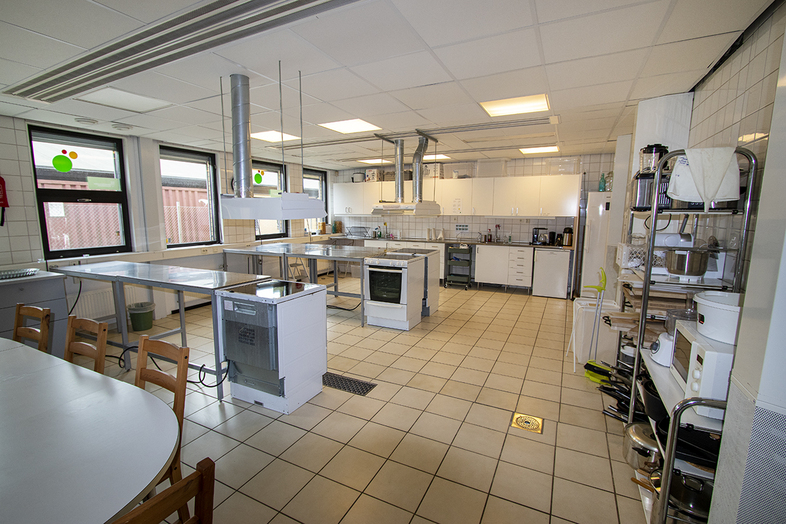 Stort, ljust storkök med flera arbetsytor, spisar, diskbänk och hyllor med köksutrustning.