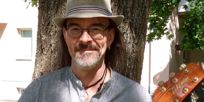 Mathias Beltramo Brandin med hatt och glasögon står framför ett träd, håller en gitarr, utomhus i dagsljus.