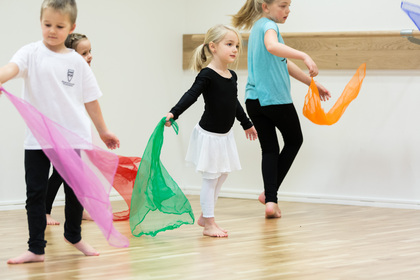 Flera barn dansar barfota i en danssal och håller färgglada tygsjalar i händerna.