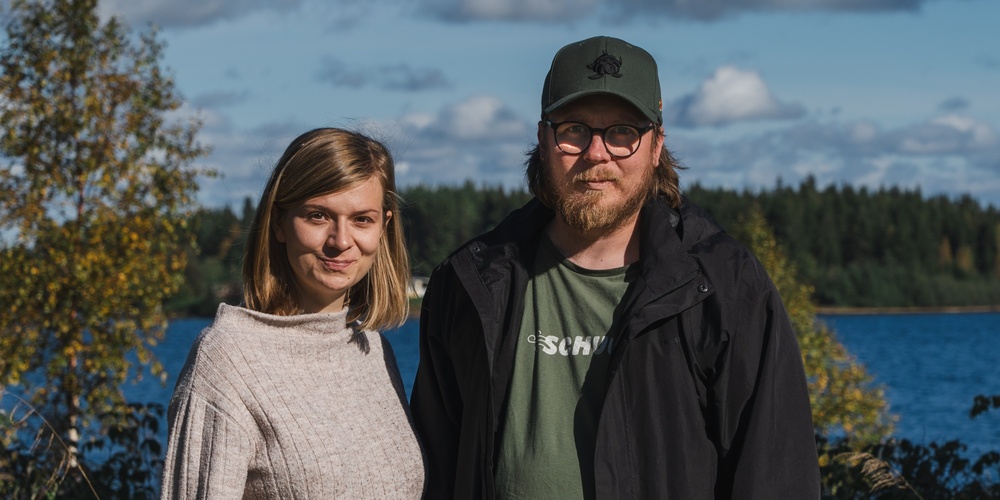 En kvinna och en man står utomhus vid en sjö med skog i bakgrunden under en solig dag.