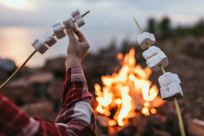En person grillar marshmallows på pinnar över en öppen eld vid en sjö eller strand.