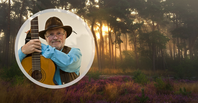 En äldre man med hatt och gitarr i en cirkel, framför en solbelyst skog med lila ljung på marken.