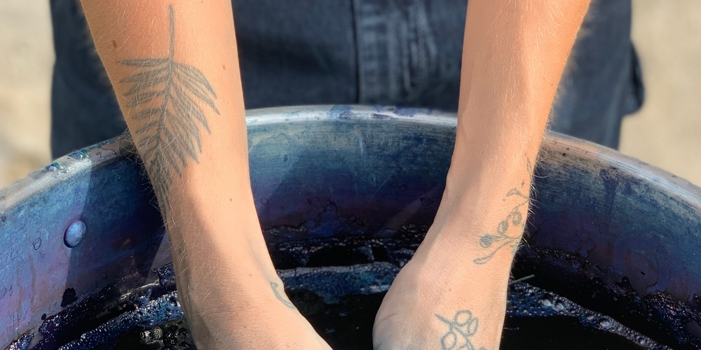 Två tatuerade armar doppas i en blå vätska i en stor behållare.