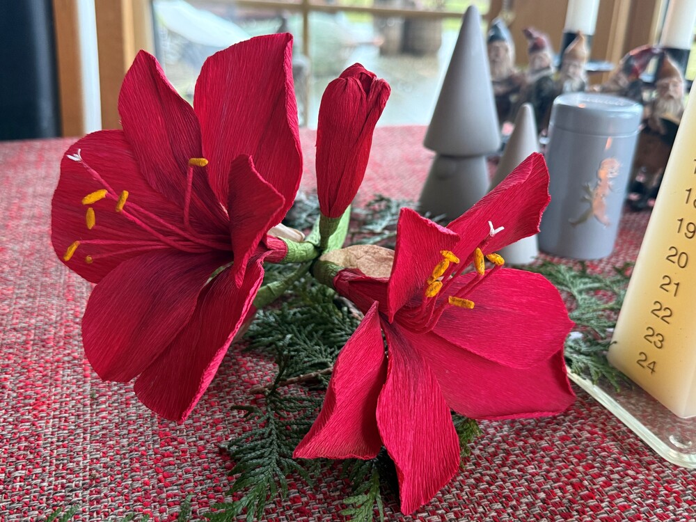Två röda amaryllisblommor av papper ligger på ett rött bord med juldekorationer i bakgrunden.