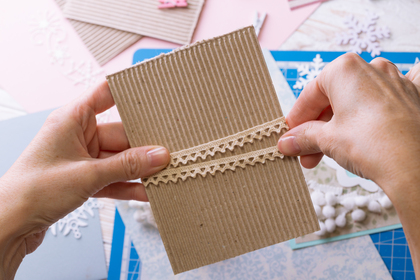 Två händer dekorerar ett kort av wellpapp med spetsband på ett pysselbord.